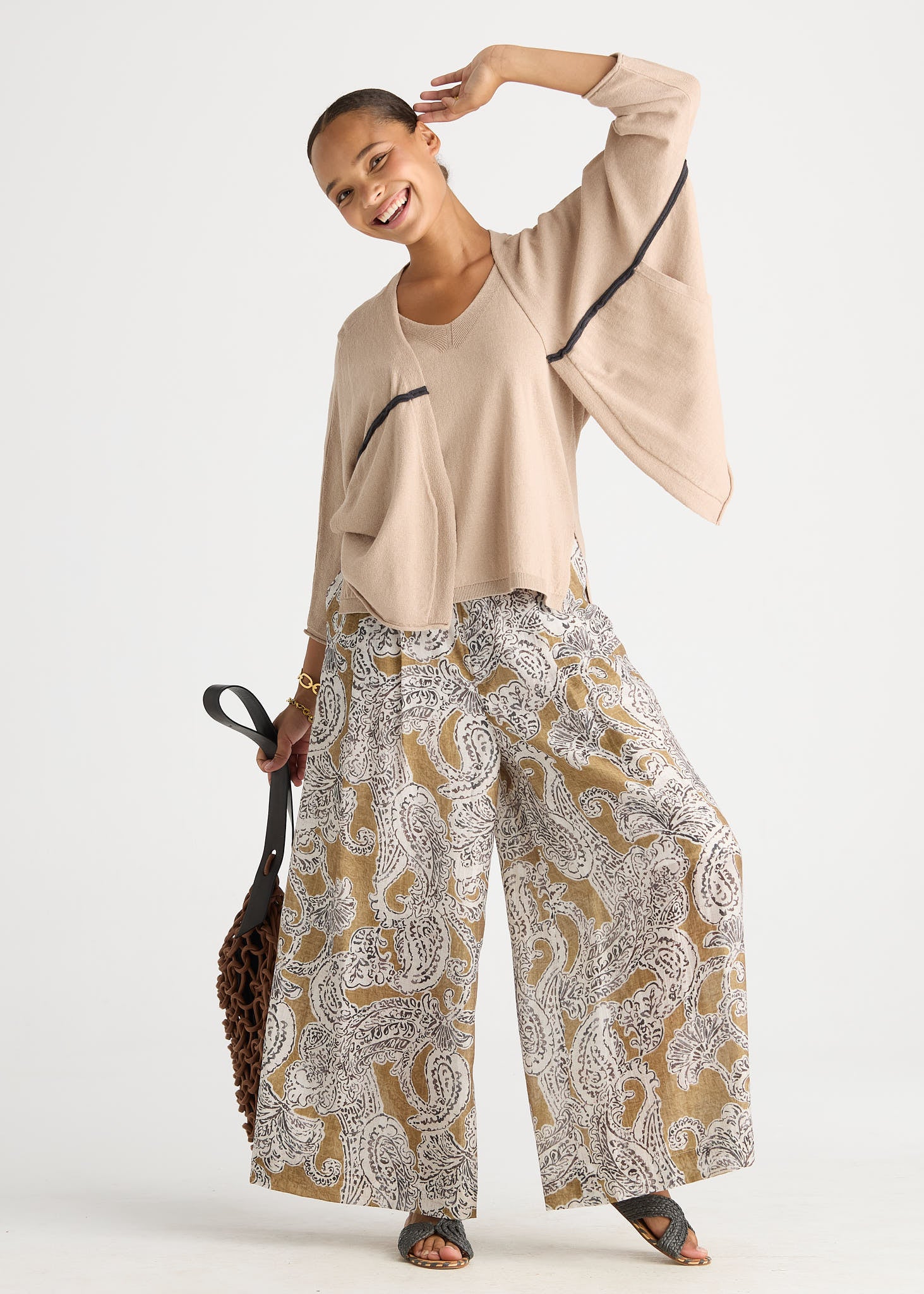 A woman modeling a beige cropped long-sleeve top with black trim and wide-leg patterned pants featuring a brown, beige, and white paisley print, holding a brown woven handbag against a light gray studio background.