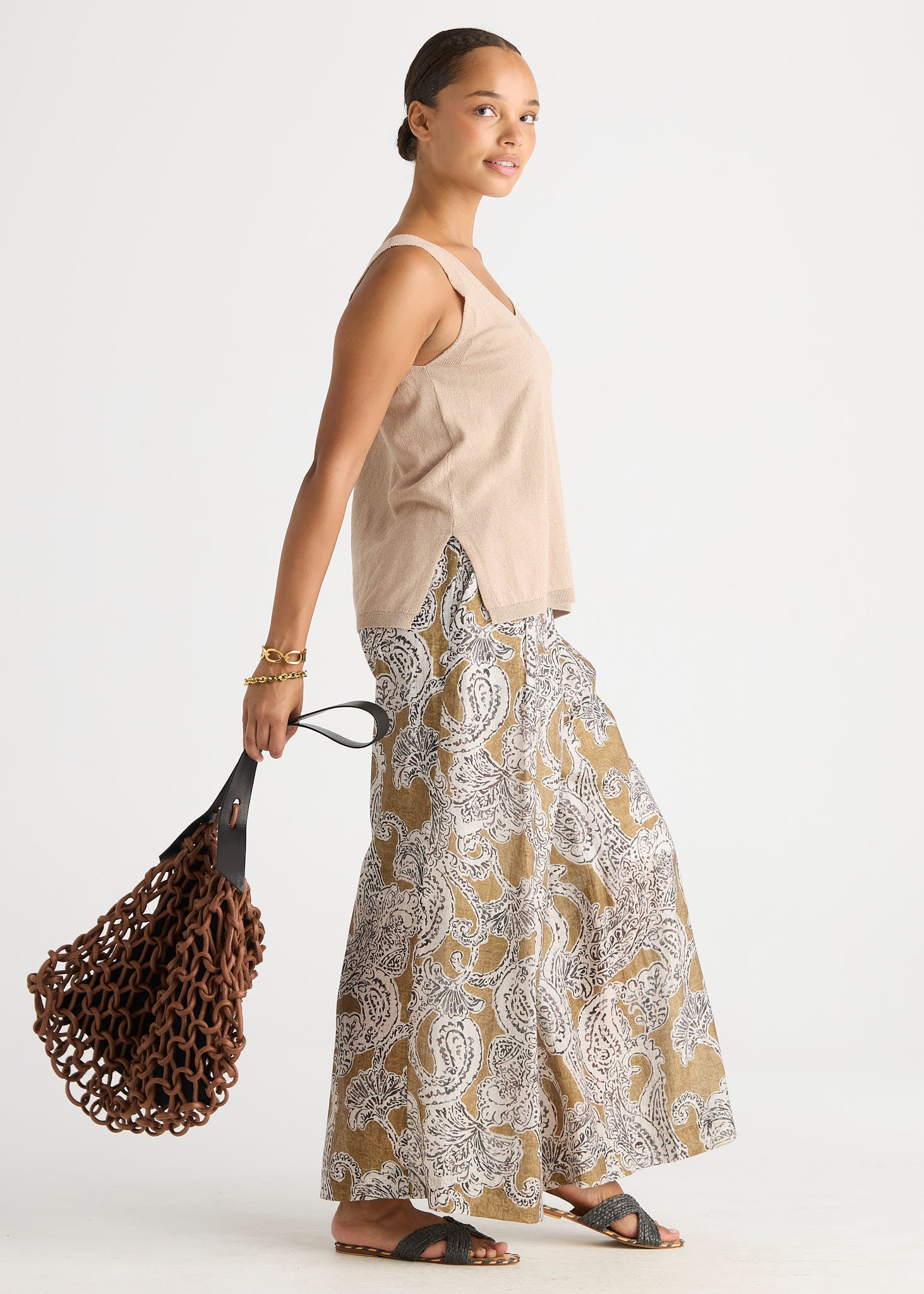 A woman modeling a sleeveless beige top and wide-leg patterned pants with a brown woven bag, photographed against a plain light gray studio background.