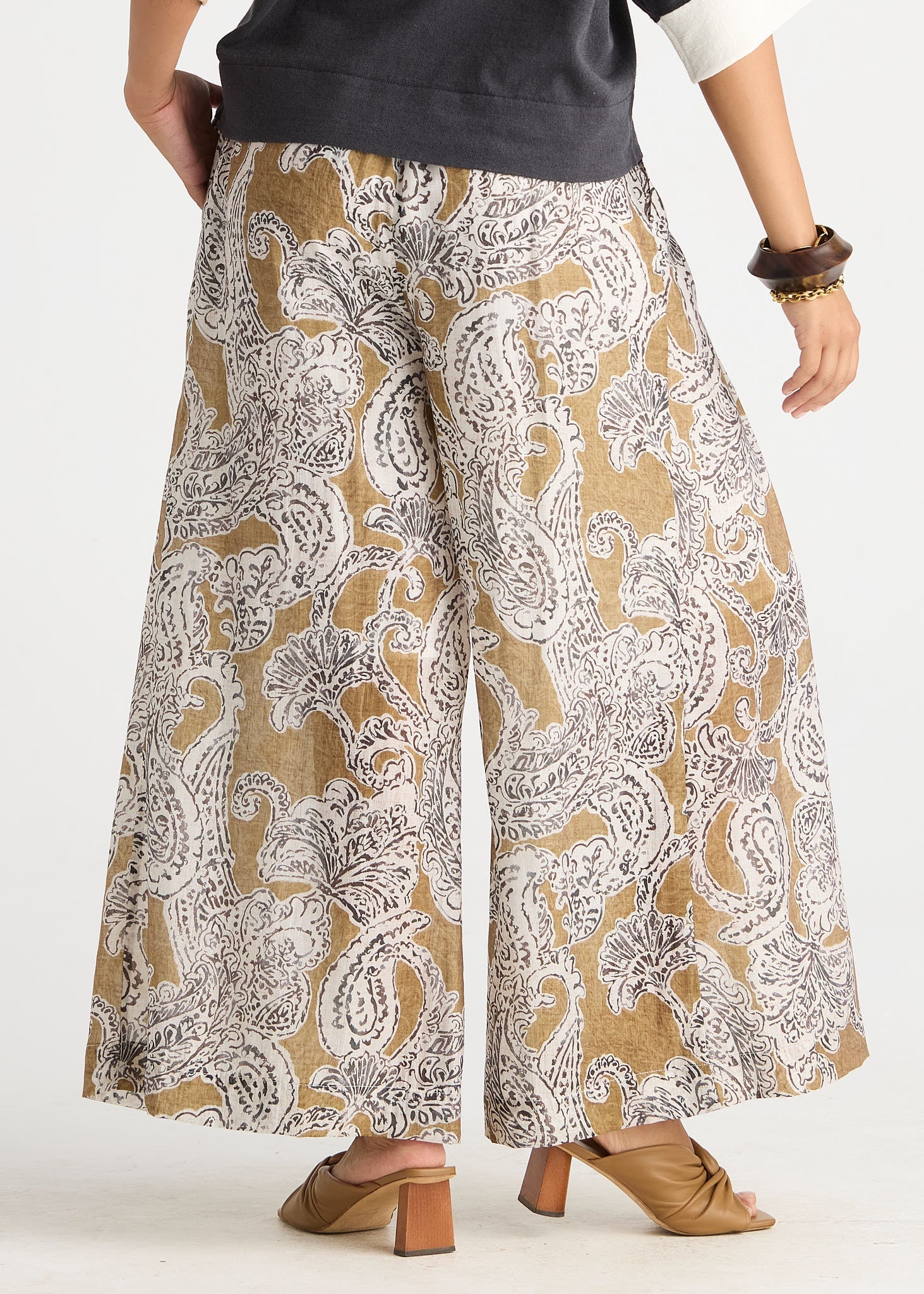 A person wearing wide-leg cropped pants with a beige, white, and gray paisley pattern, styled with a dark gray top and tan heeled mule sandals against a plain white studio background.
