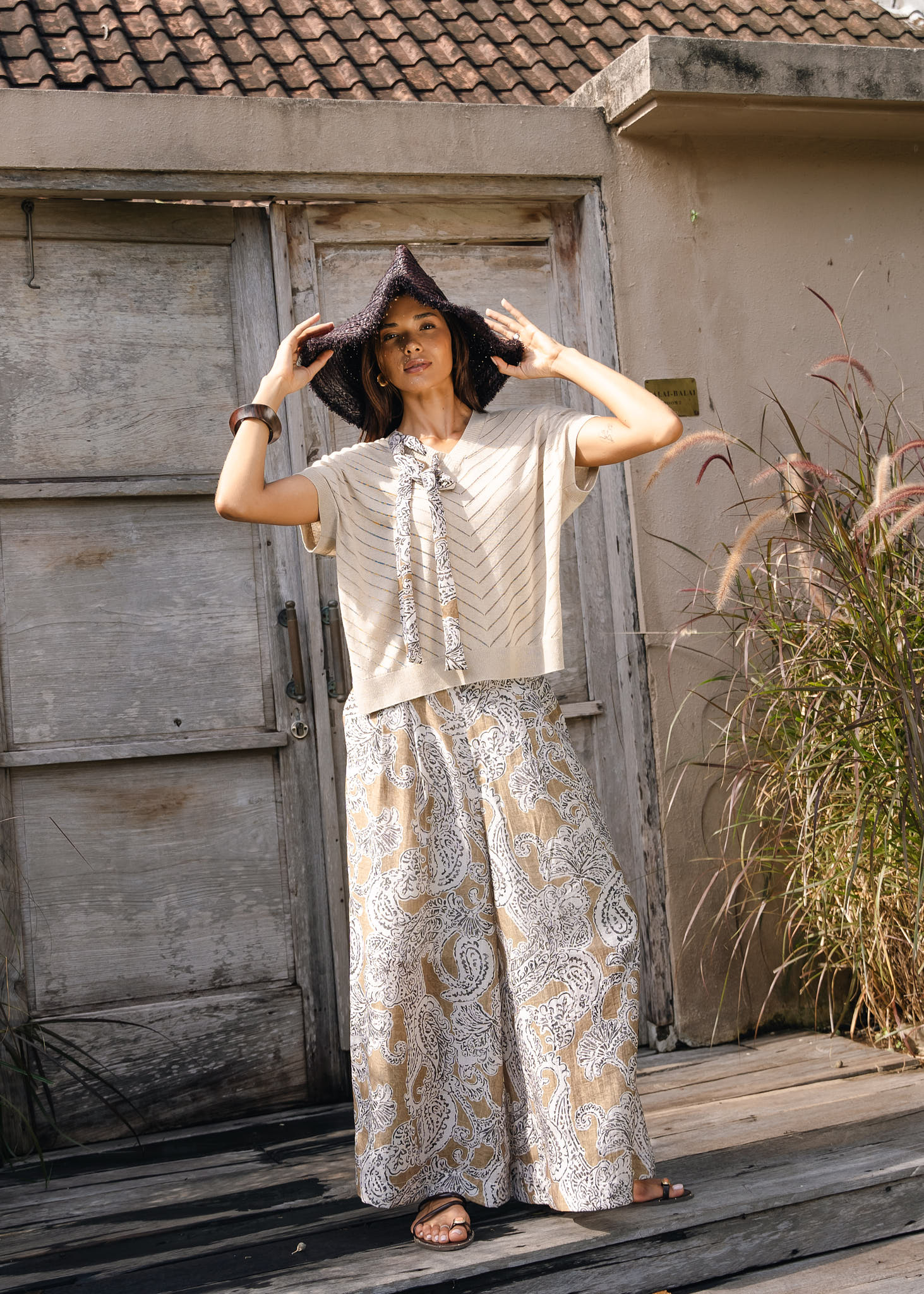 A woman stands on a wooden deck in front of a rustic weathered door and wall, wearing a beige short-sleeve top with a tie detail, matching beige and white paisley wide-leg pants, and a dark wide-brim hat.