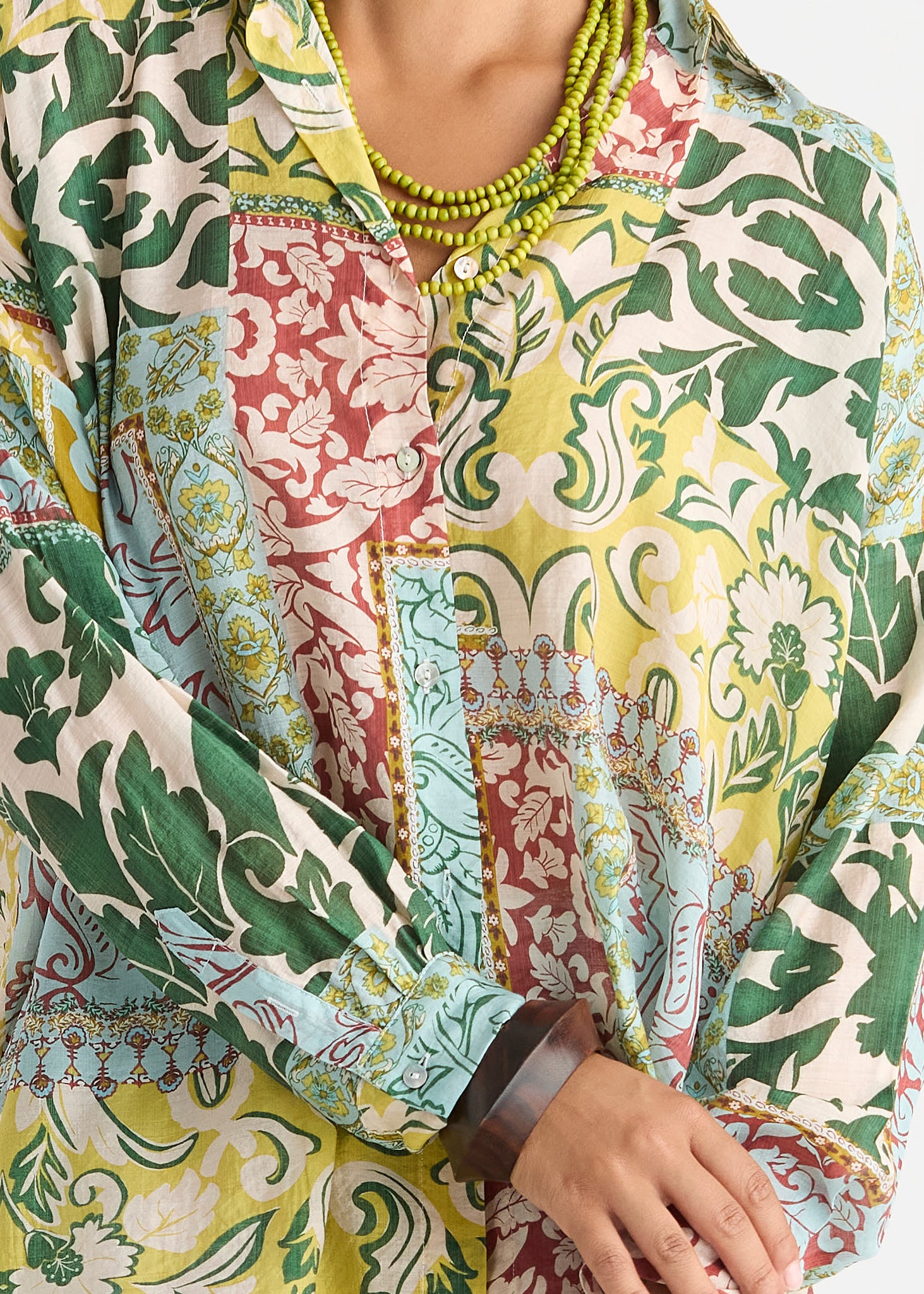 A close-up of a person wearing a long-sleeve button-up shirt with a vibrant multicolor floral and paisley print in green, yellow, red, and blue, accessorized with a chunky green beaded necklace and a brown cuff bracelet.