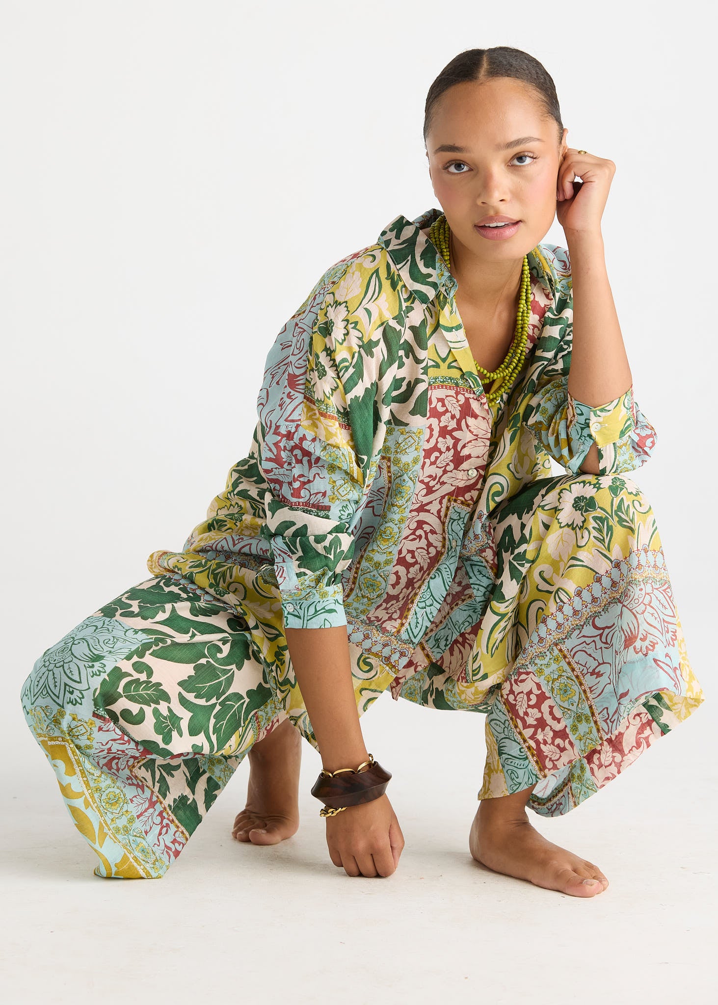 A woman crouches barefoot on a white studio floor wearing a loose, patterned kaftan-style dress with green, yellow, red, and blue botanical motifs.