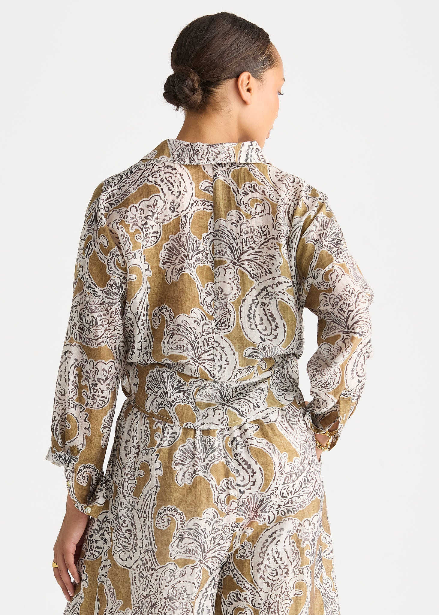 A woman shown from the back wearing a long-sleeve belted jumpsuit with a brown, beige, and white paisley pattern, set against a plain light gray background.