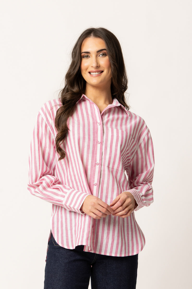 A woman with long dark hair wearing a pink and white vertically striped button-down shirt and dark jeans, standing against a plain off-white studio background.