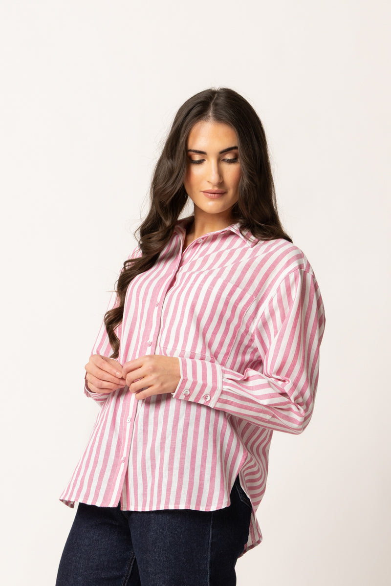 A woman wearing a pink and white vertically striped button-down shirt with dark blue jeans, standing against a plain off-white studio background.