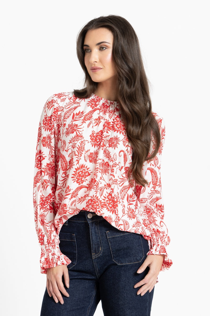 A woman modeling a long-sleeve blouse with a red and orange floral paisley pattern on a white background, paired with dark blue jeans, against a light gray studio background.