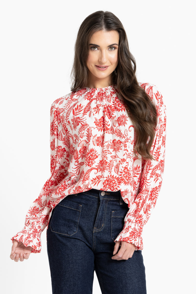 A woman modeling a long-sleeve blouse with a red and white floral pattern, paired with dark blue jeans, against a plain white background.
