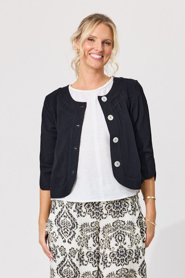 A smiling blonde woman models a short black cropped jacket with silver buttons over a white top and black-and-white patterned pants, shot against a light gray studio background.