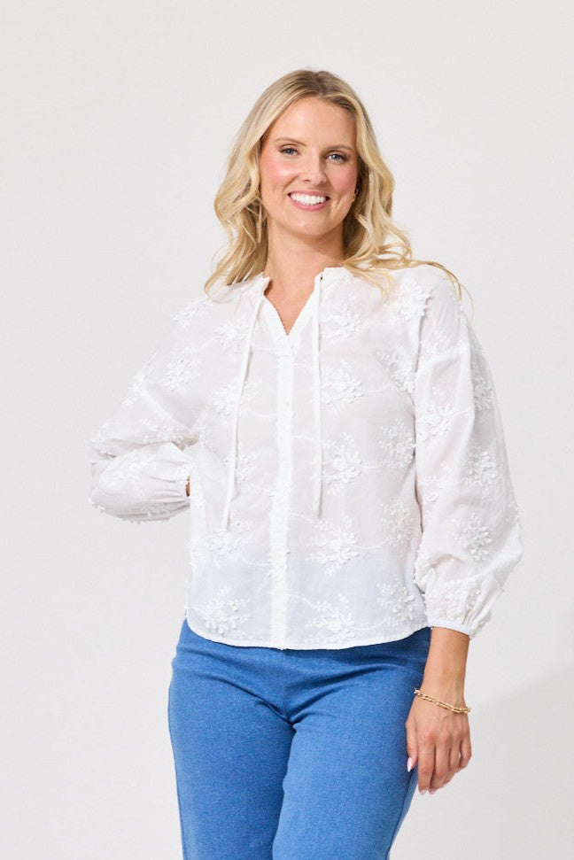 A smiling blonde woman wearing a white long-sleeve blouse with floral embroidery and drawstrings, paired with blue jeans, against a plain light gray studio background.
