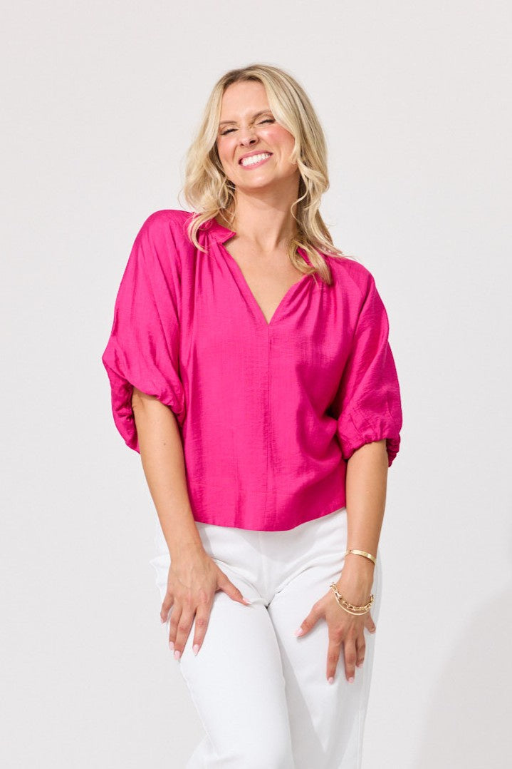 A smiling blonde woman wearing a bright fuchsia pink blouse with voluminous elbow-length puff sleeves and white pants, standing against a light gray studio background.