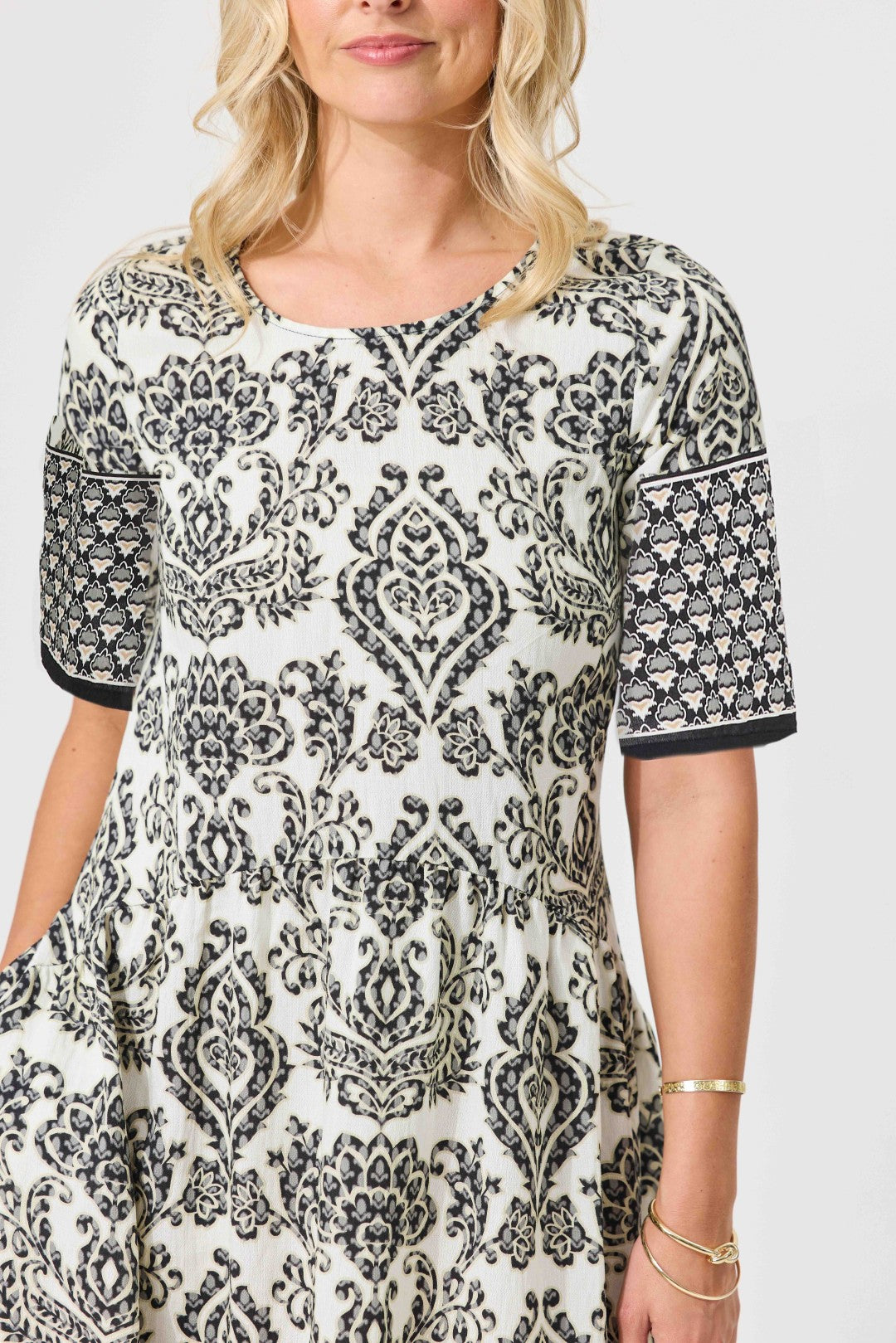 A woman wearing a short-sleeve tunic dress with a black and white damask pattern, featuring solid black cuffs and a scoop neckline, photographed against a light gray studio background.