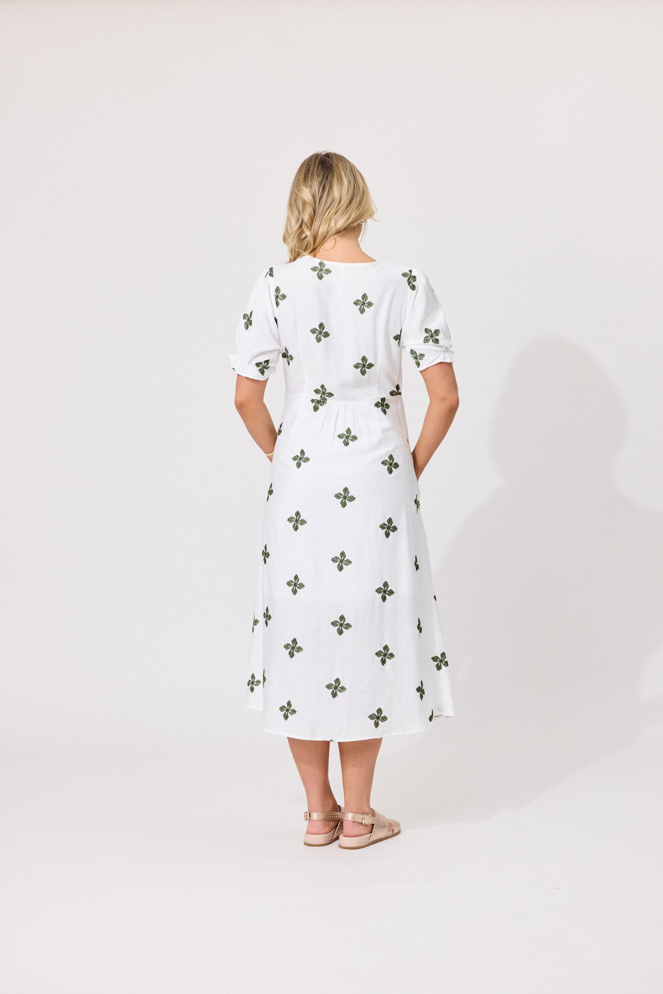 A woman viewed from behind wearing a white midi dress with short puff sleeves and an allover green clover pattern, standing against a plain light gray studio background.