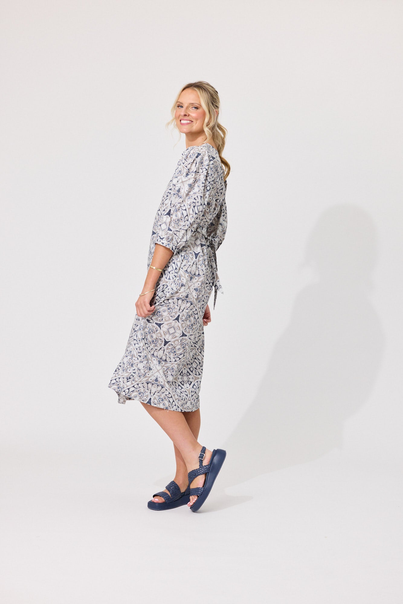 A woman modeling a knee-length wrap dress with a gray and white floral pattern, paired with navy blue slide sandals, standing against a plain light gray studio background.