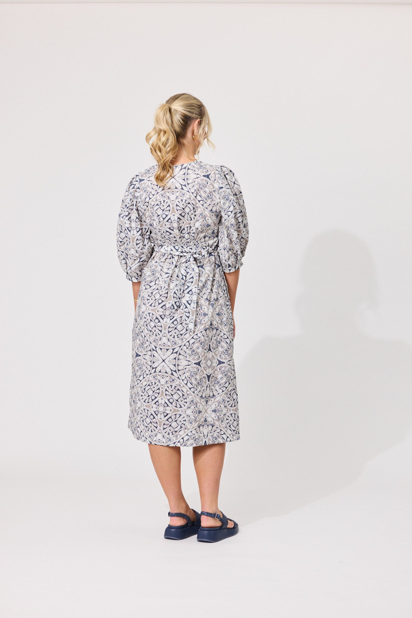 A woman viewed from behind wearing a knee-length patterned dress with puff sleeves and a tie waist, paired with dark blue Mary Jane sandals, against a plain white studio background.