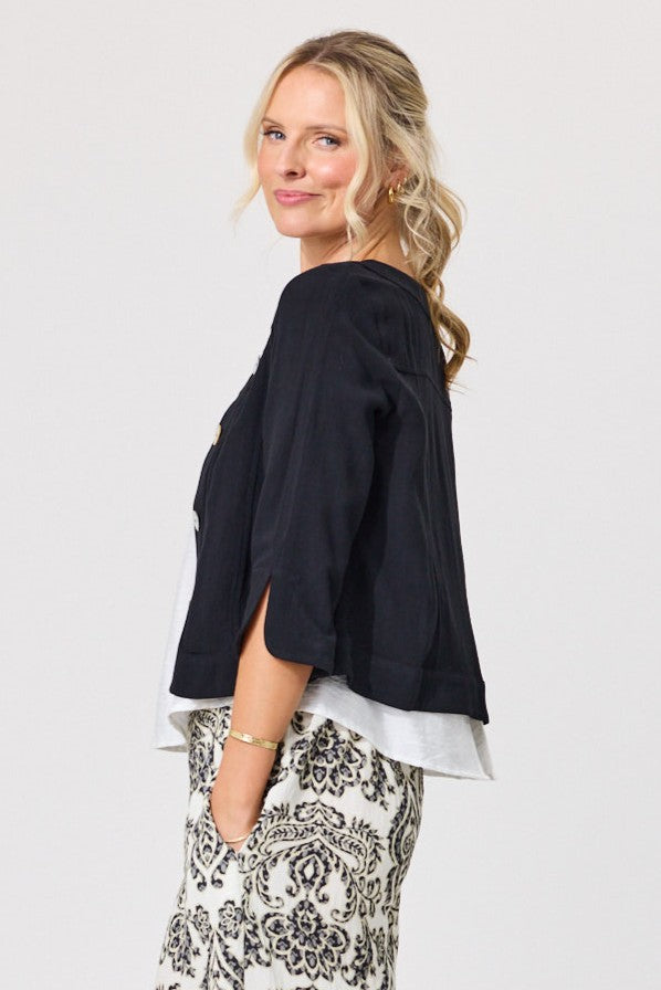 A woman modeling a black cropped top with a layered white underlayer and black-and-white patterned pants, photographed against a plain light gray studio background.