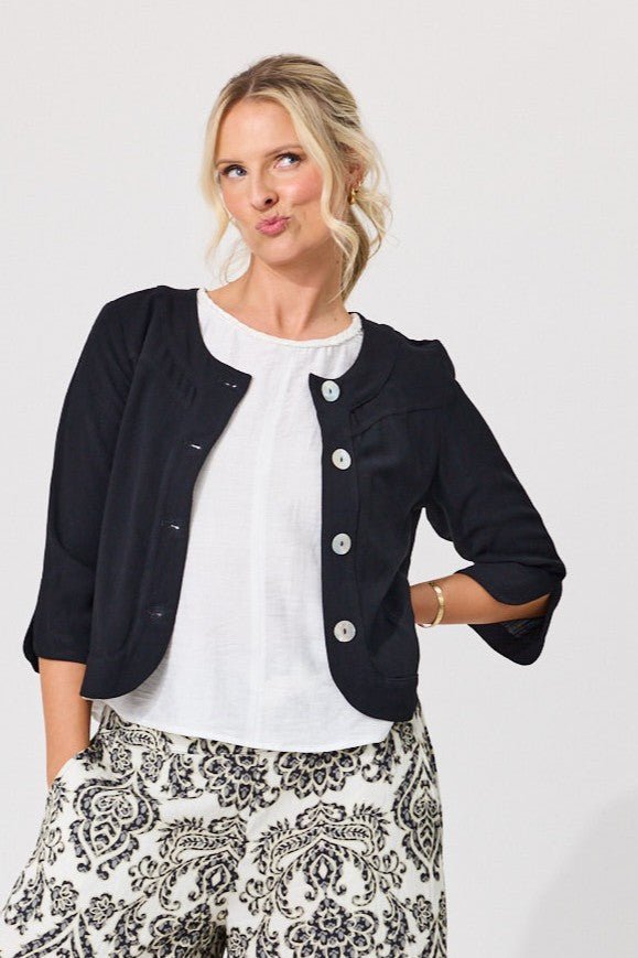 A woman modeling a black cropped jacket with three-quarter sleeves and decorative silver buttons, worn over a white top and black-and-white patterned pants, against a plain light gray background.