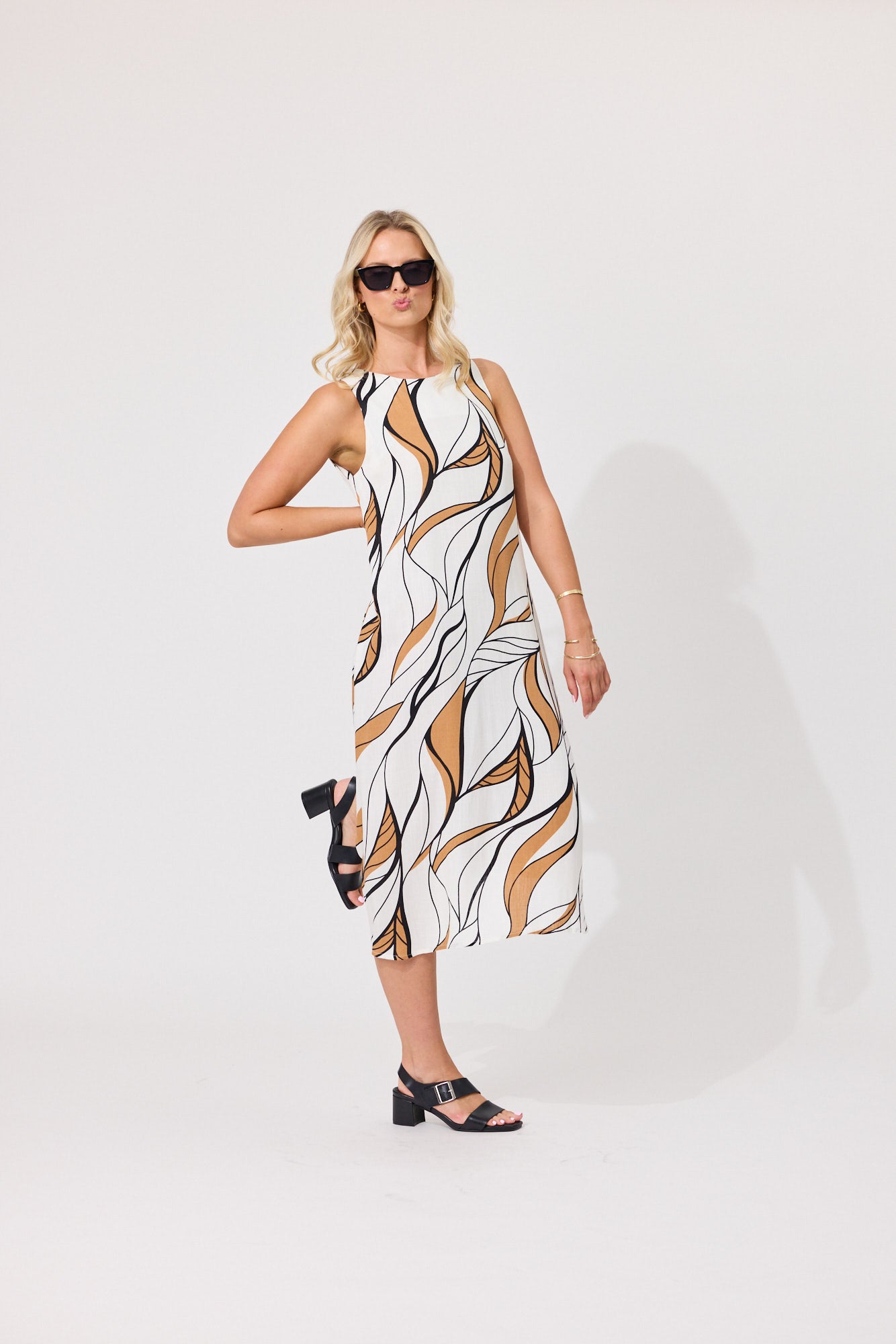A woman modeling a sleeveless midi dress with an abstract wavy pattern in white, black, and tan, styled with black strappy heels and sunglasses against a plain white studio background.
