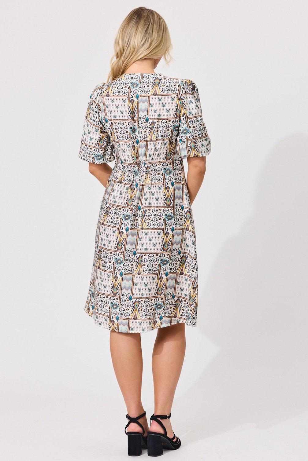 A woman viewed from the back wearing a knee-length dress with puff sleeves and an allover patchwork-style print in beige, white, blue, and brown tones. She is standing against a plain light gray studio background and wearing black high-heeled sandals.