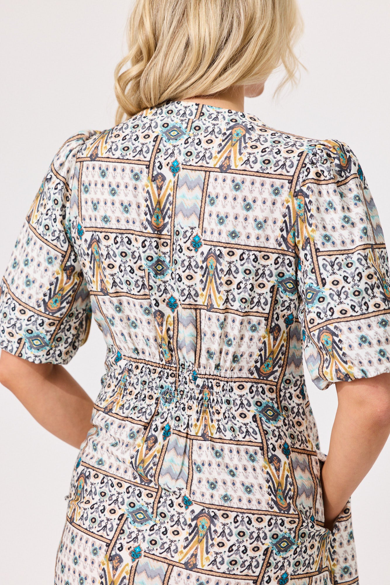 A woman shown from the back wearing a short-sleeve dress with a detailed geometric and tribal-inspired print in blue, yellow, brown, and black on a cream background.
