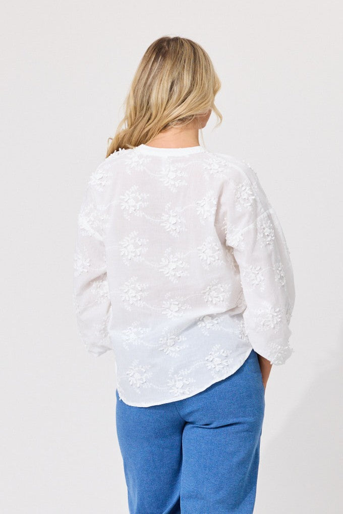 A woman viewed from the back wearing a white long-sleeve blouse with delicate floral embroidery and blue jeans, set against a plain light gray studio background.
