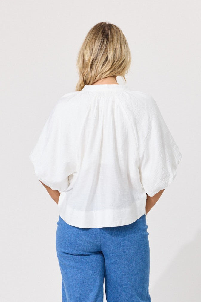 A woman viewed from behind wearing a white short-sleeve blouse with voluminous puff sleeves and blue pants, set against a plain light gray studio background.