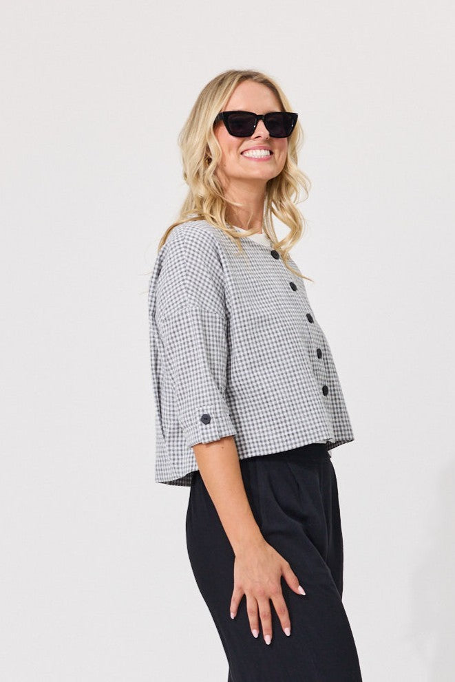 A woman modeling a cropped black and white gingham double-breasted jacket with three-quarter sleeves and black buttons, paired with black trousers and sunglasses, against a plain light gray background.