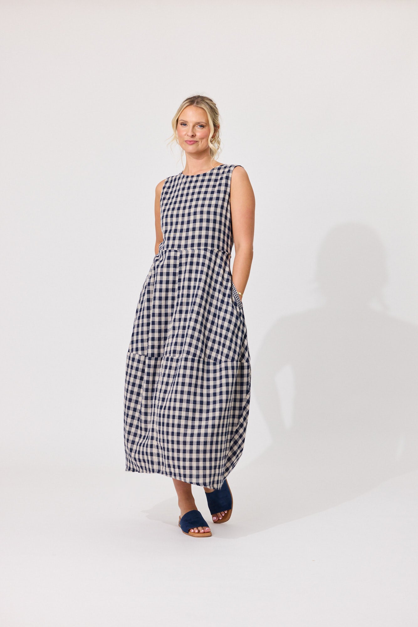 A woman modeling a sleeveless black and white gingham maxi dress with pockets, standing against a plain light gray studio background.