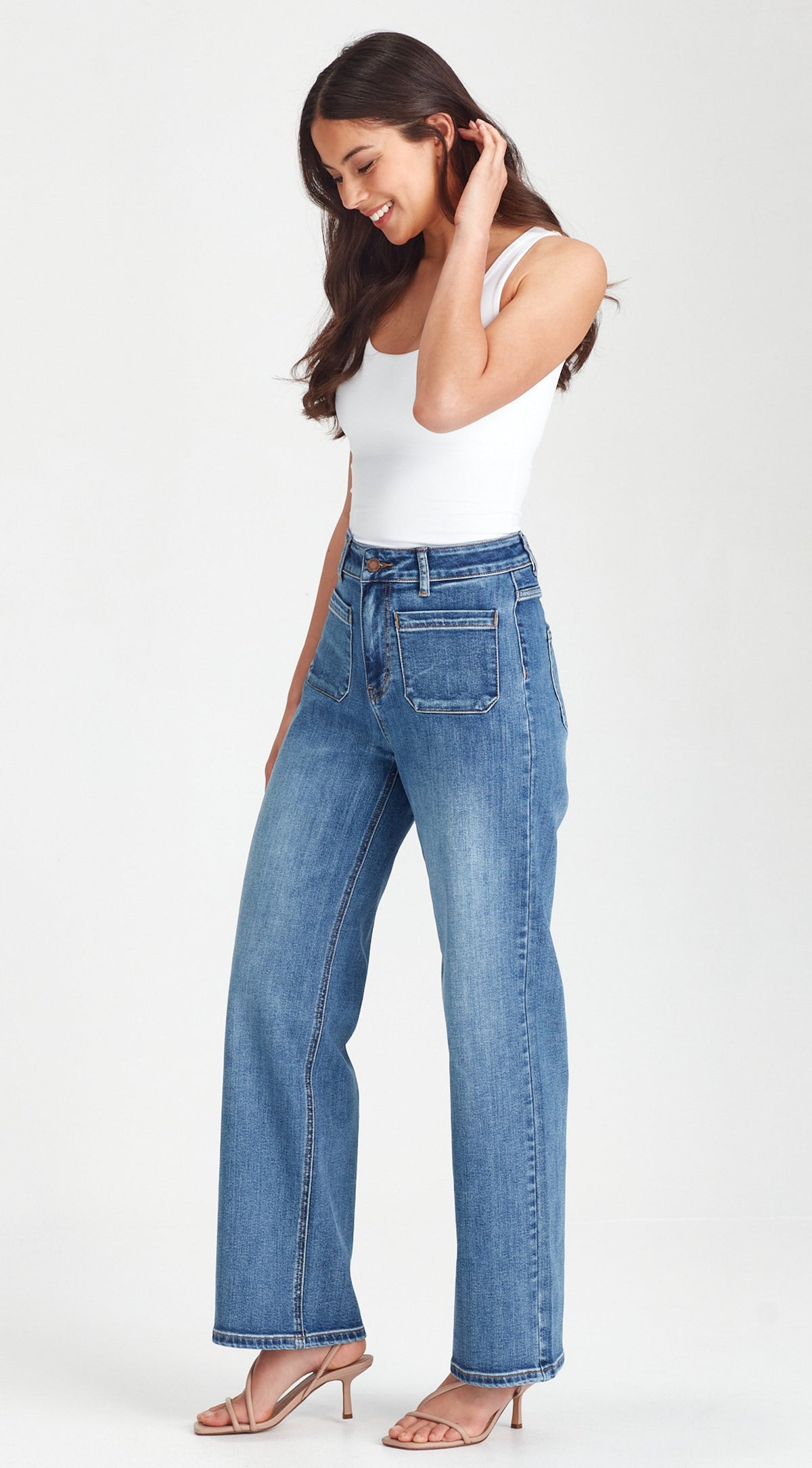 A woman modeling a pair of high-waisted, wide-leg blue jeans with a white tank top and beige heeled sandals, standing against a plain white studio background.