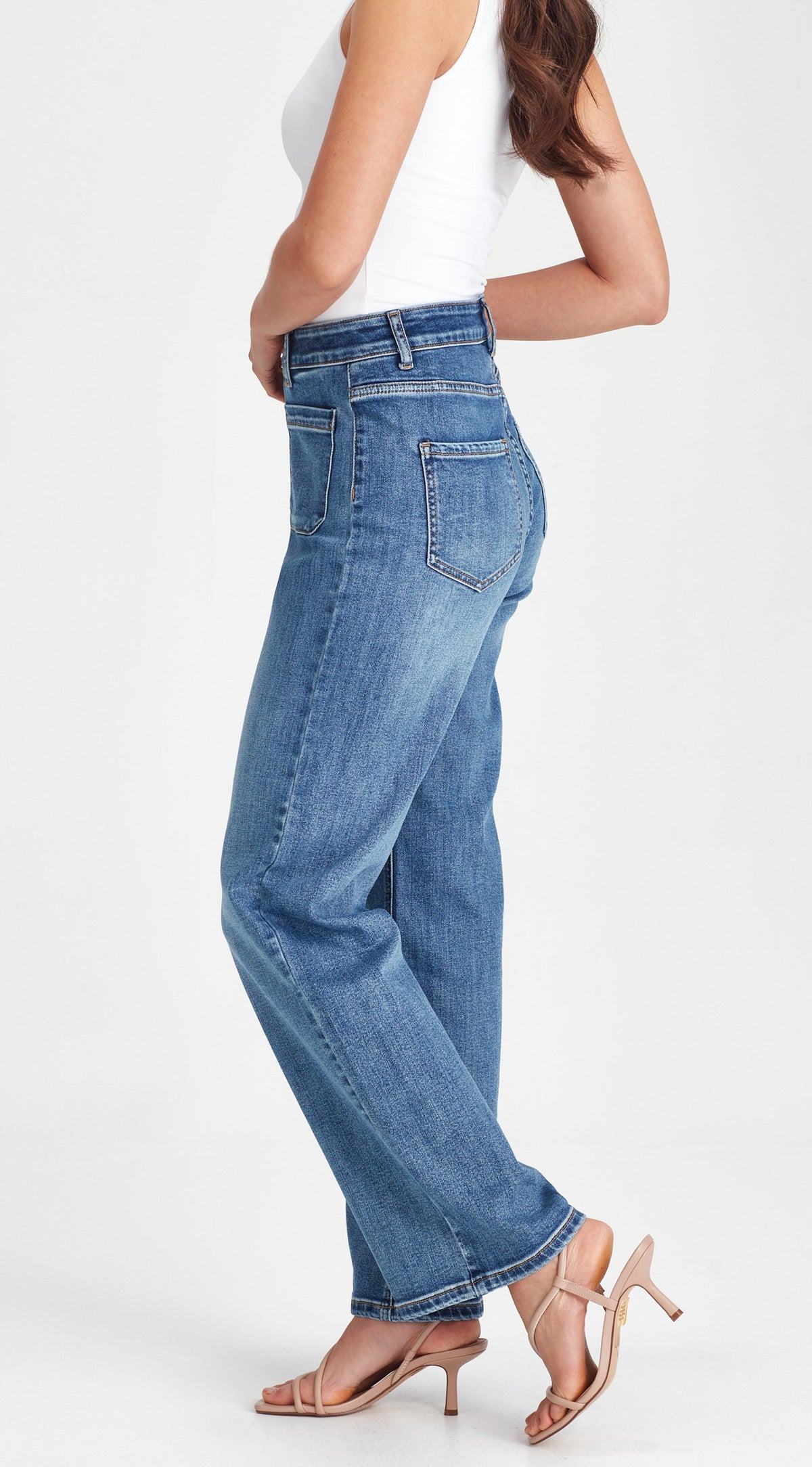 A woman wearing high-waisted, wide-leg blue denim jeans, a white sleeveless top, and beige high-heeled sandals, shown from a side-back angle against a plain white studio background.