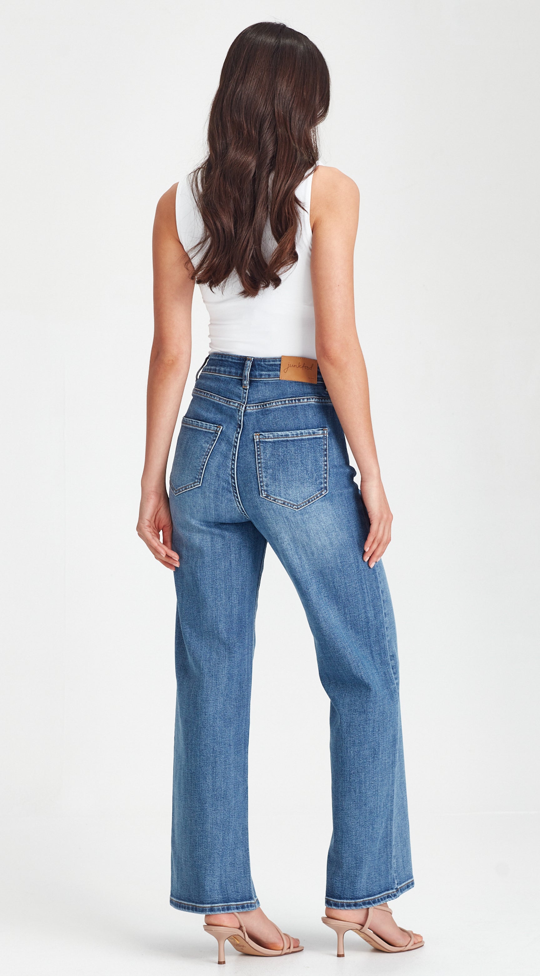 A woman viewed from the back wearing blue wide-leg jeans, a white sleeveless top, and nude high-heeled sandals against a light gray studio background.