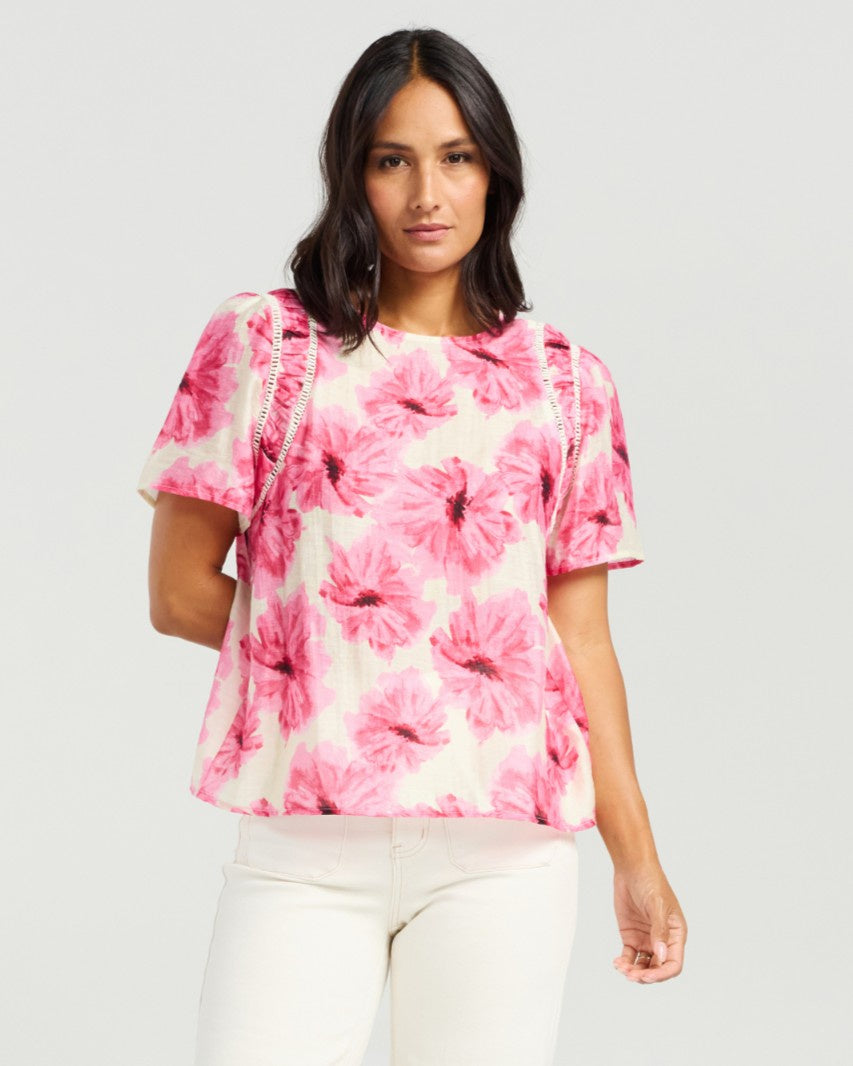 A woman modeling a short-sleeve blouse with a pink and white floral print, paired with white pants, against a light gray studio background.