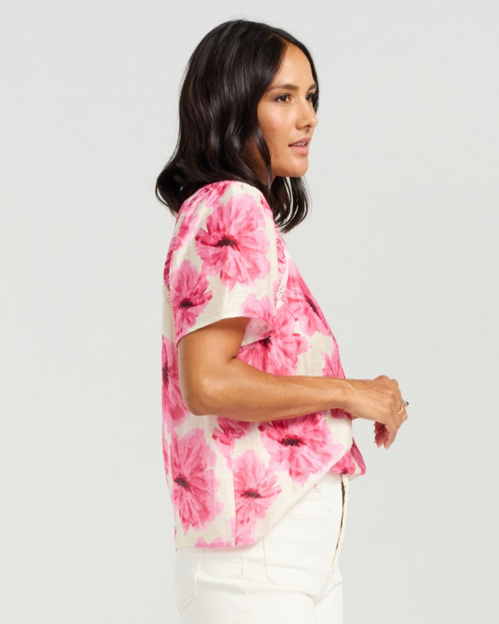 A woman shown in side profile wearing a short-sleeve blouse with a pink and magenta floral print on a white base, paired with white pants, against a light gray studio background.