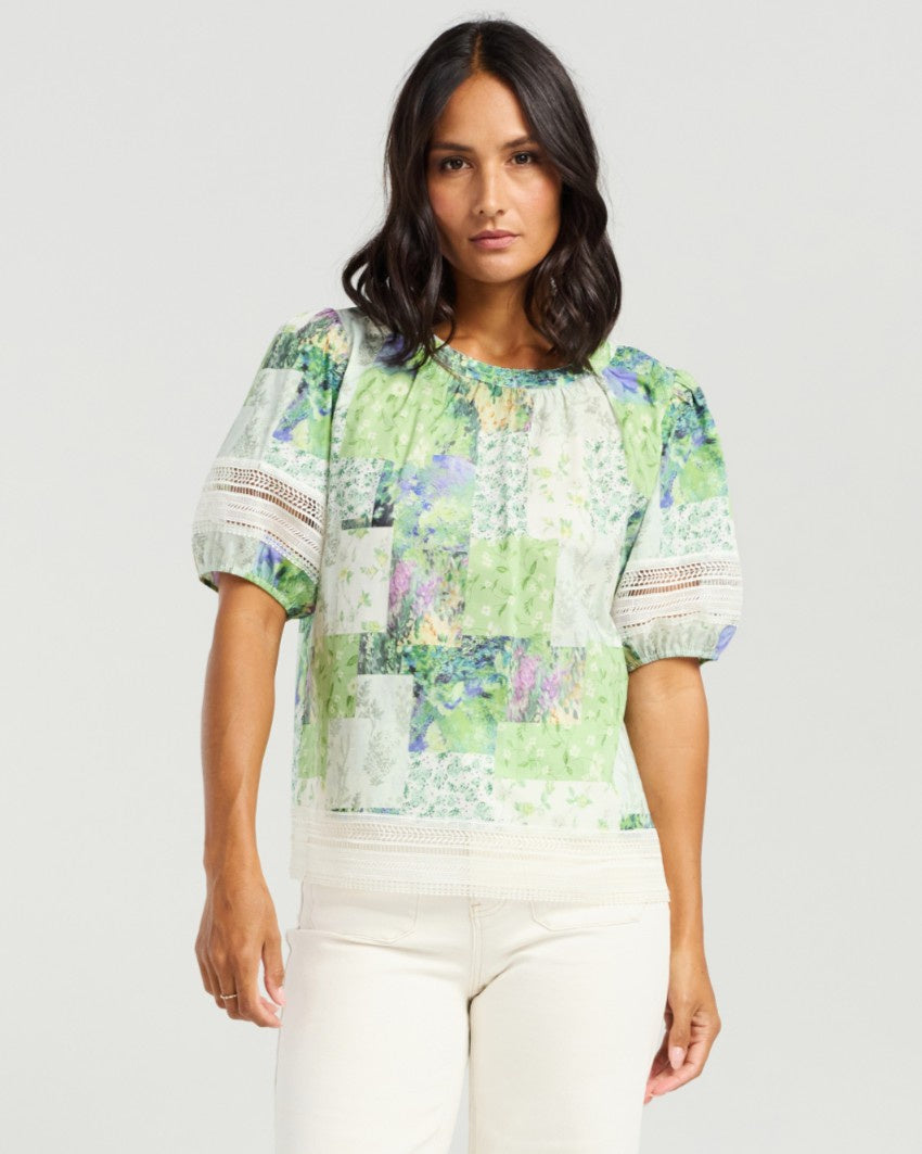 A woman modeling a short-sleeve patchwork blouse with green, white, and light blue floral prints, paired with off-white pants, against a light gray studio background.
