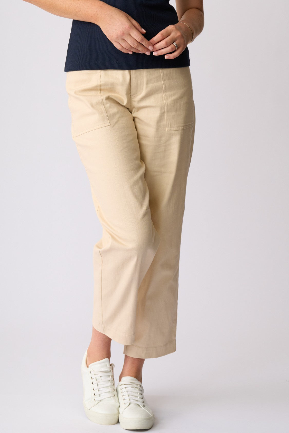 A person wearing light beige cropped pants, a dark navy top, and white sneakers, standing against a plain light gray studio background.