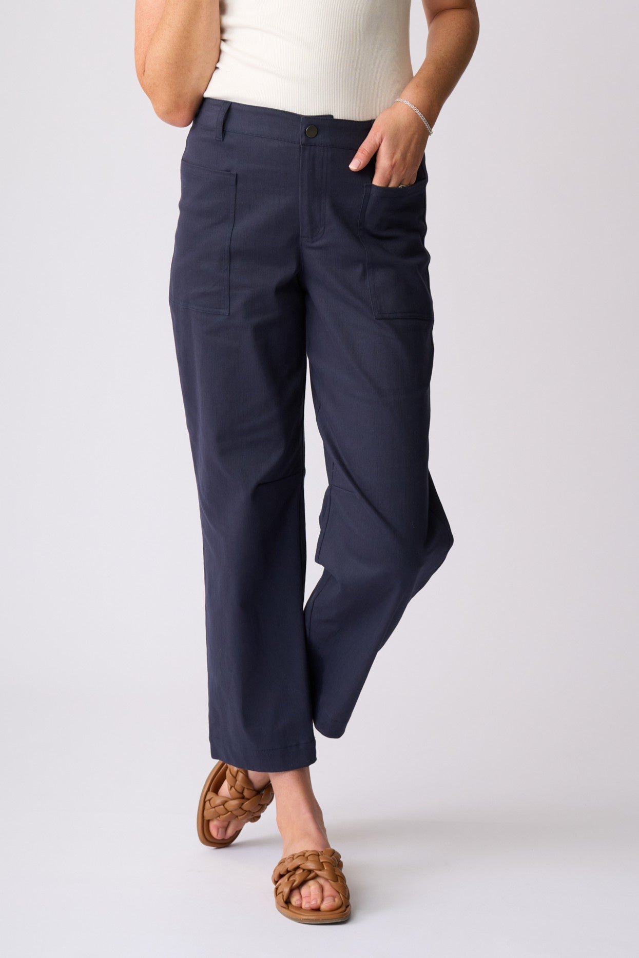 A person wearing dark navy cropped pants with a white top and brown woven slide sandals, standing against a light gray studio background.