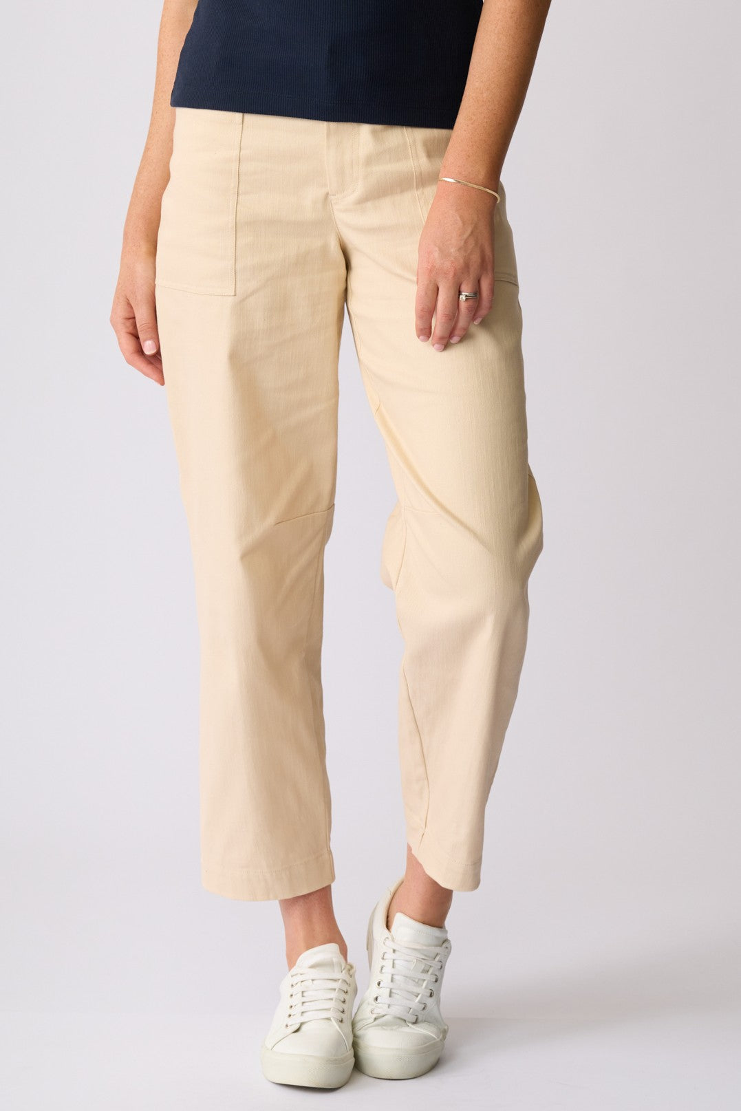 A person wearing light beige cropped trousers with a navy top and white sneakers, standing against a light gray studio background.