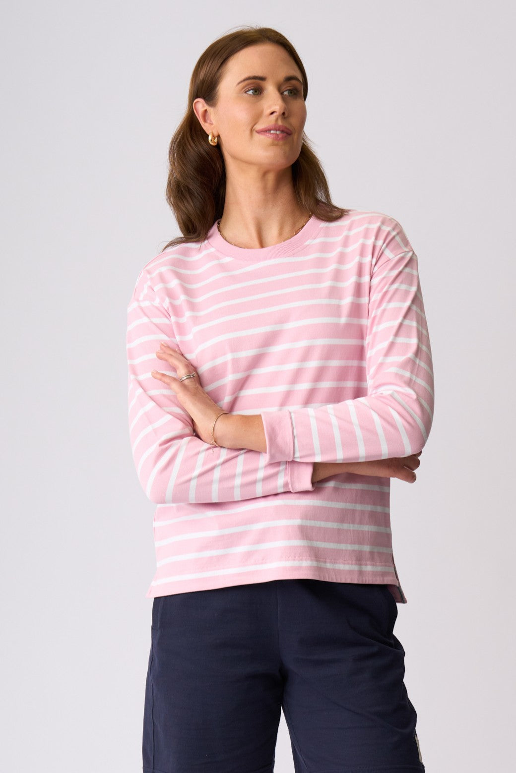 A woman modeling a pink and white striped long-sleeve crewneck top with dark navy pants, standing against a light gray studio background.