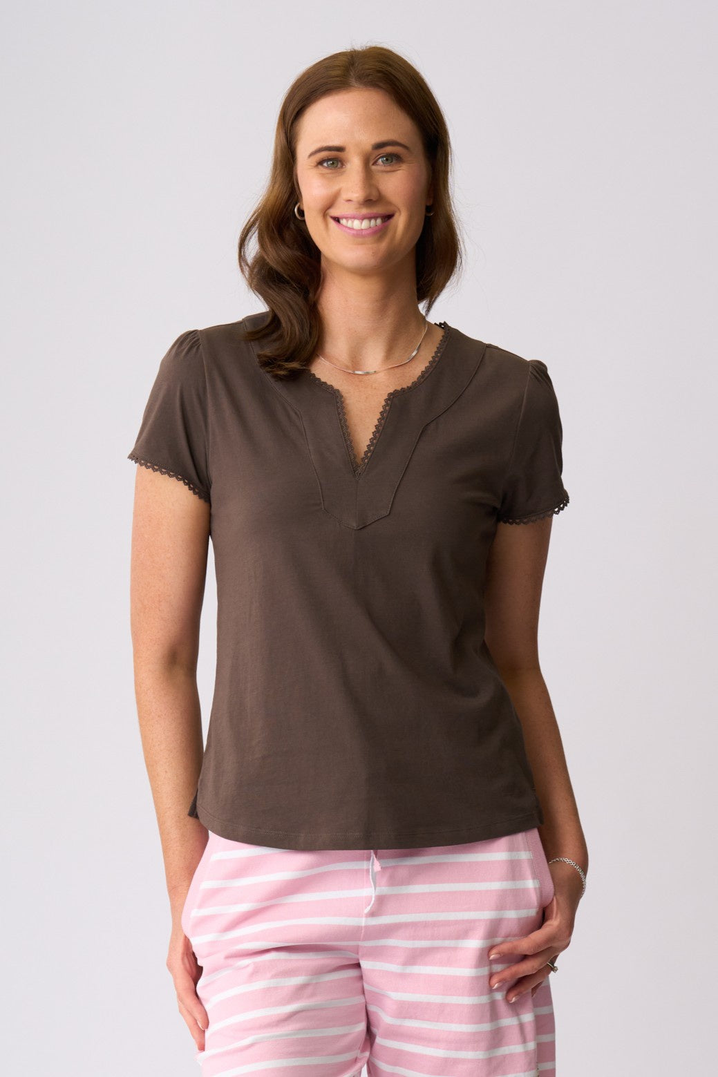 A woman modeling a brown short-sleeve V-neck top with scalloped trim, paired with pink and white striped pants, against a light gray studio background.
