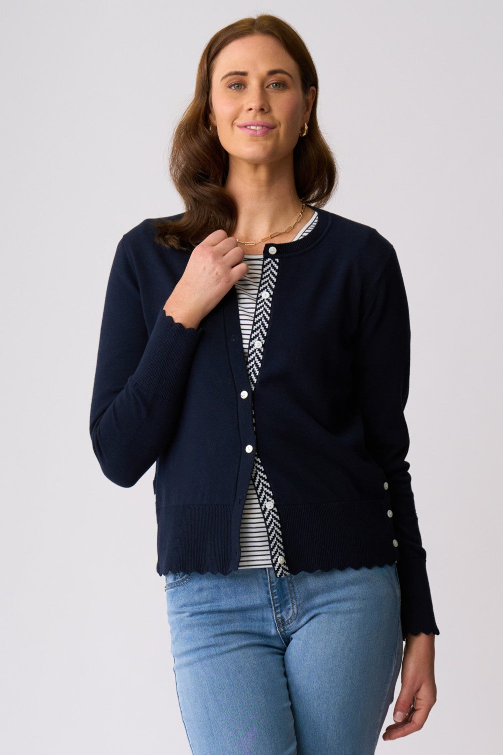 A woman modeling a navy blue cardigan with a black and white chevron patterned placket, worn over a matching striped shirt and blue jeans, against a light gray studio background.