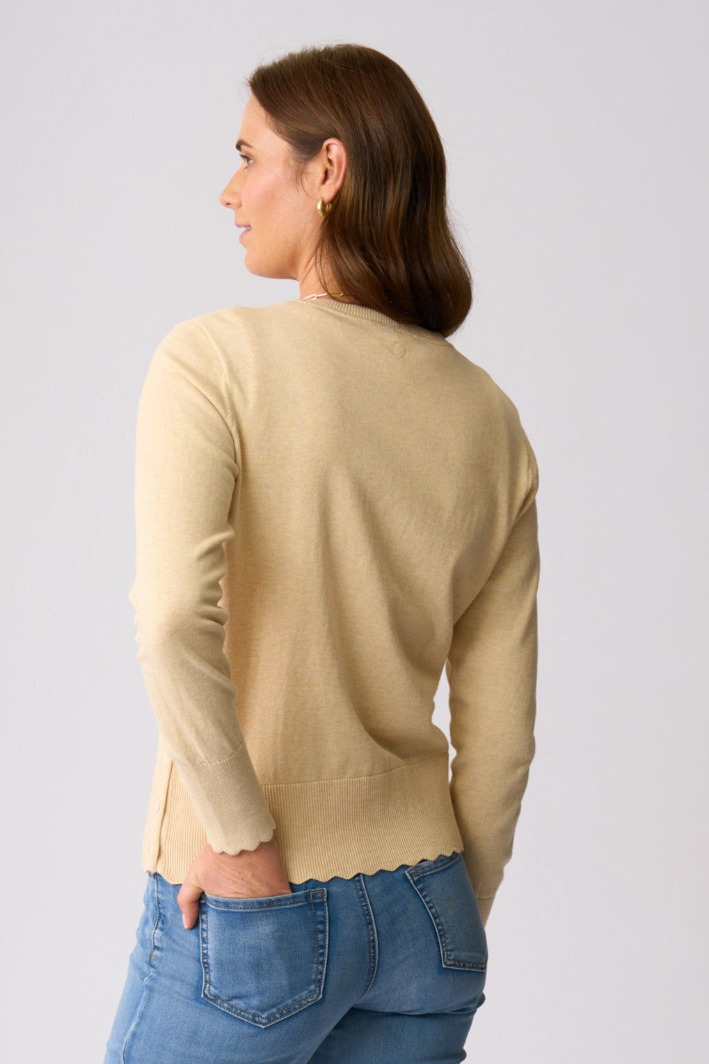 A woman shown from the back wearing a light beige knit sweater with a scalloped hem and long sleeves, paired with blue jeans, against a solid light gray background.