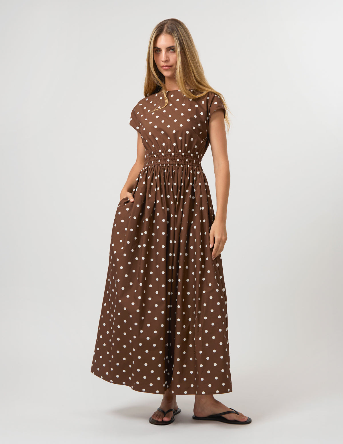 A woman modeling a brown short-sleeve maxi dress with a white polka dot pattern, standing against a light gray studio background.