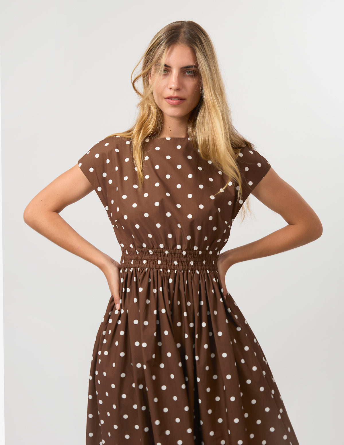 A woman modeling a brown short-sleeve dress with white polka dots, featuring a gathered elastic waist, against a plain light gray studio background.