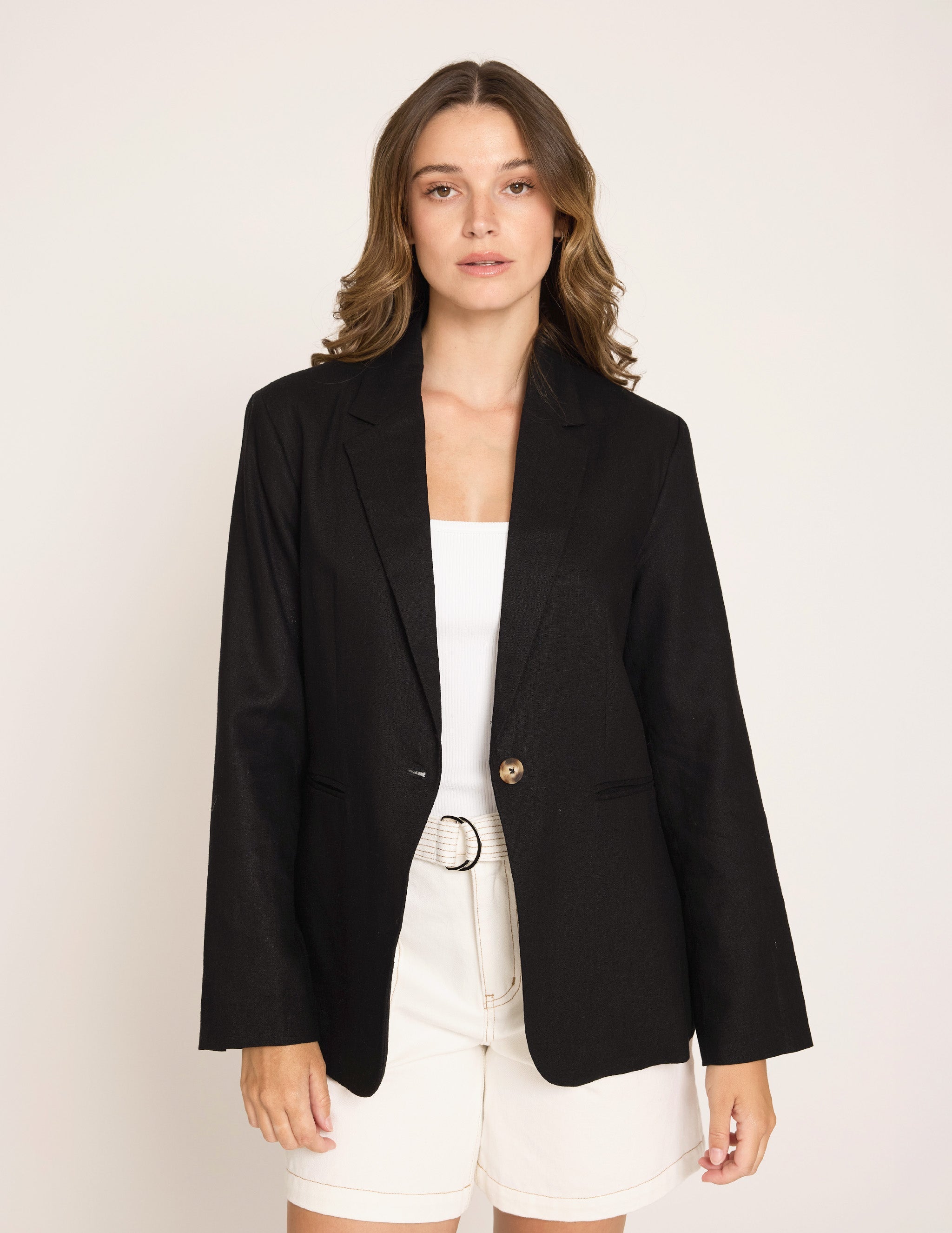A woman modeling a black single-breasted blazer with gold buttons, worn over a white top and white shorts, against a plain light gray studio background.