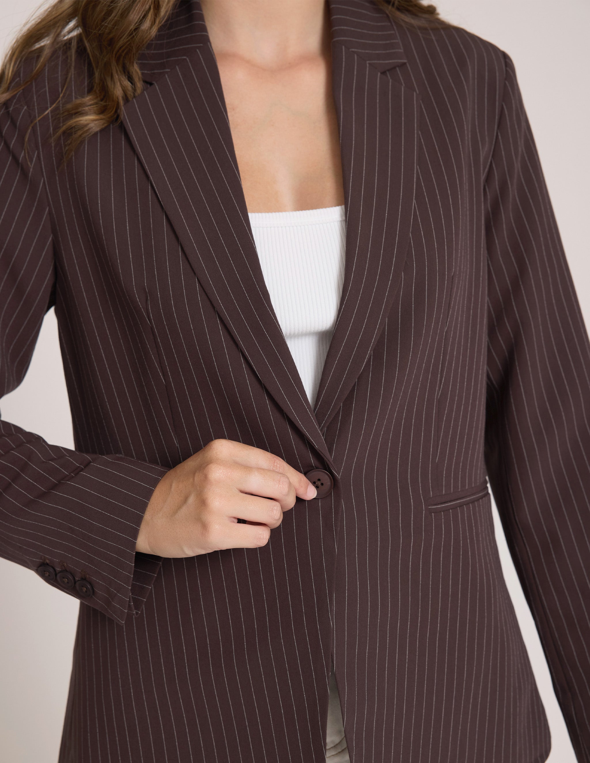 A close-up of a woman wearing a dark brown pinstripe blazer over a white ribbed tank top, with her hand adjusting the front button.