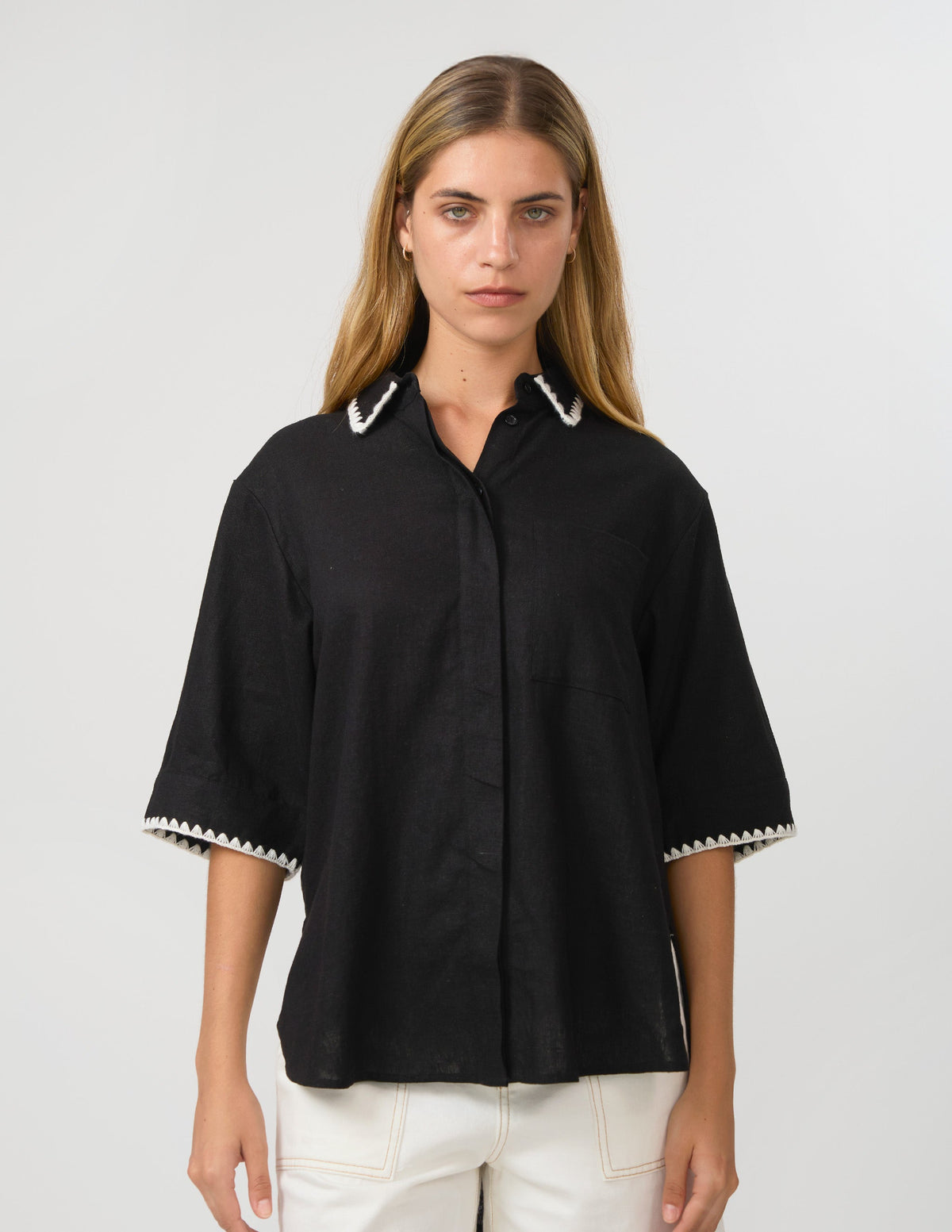 A woman modeling a black short-sleeve button-up shirt with white contrast trim on the collar and sleeve cuffs, paired with white pants, against a light gray studio background.