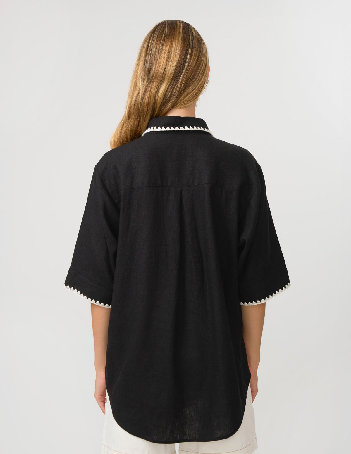 A woman viewed from the back wearing a black short-sleeve shirt with white scalloped trim on the collar and sleeve cuffs, paired with white bottoms, against a light gray studio background.