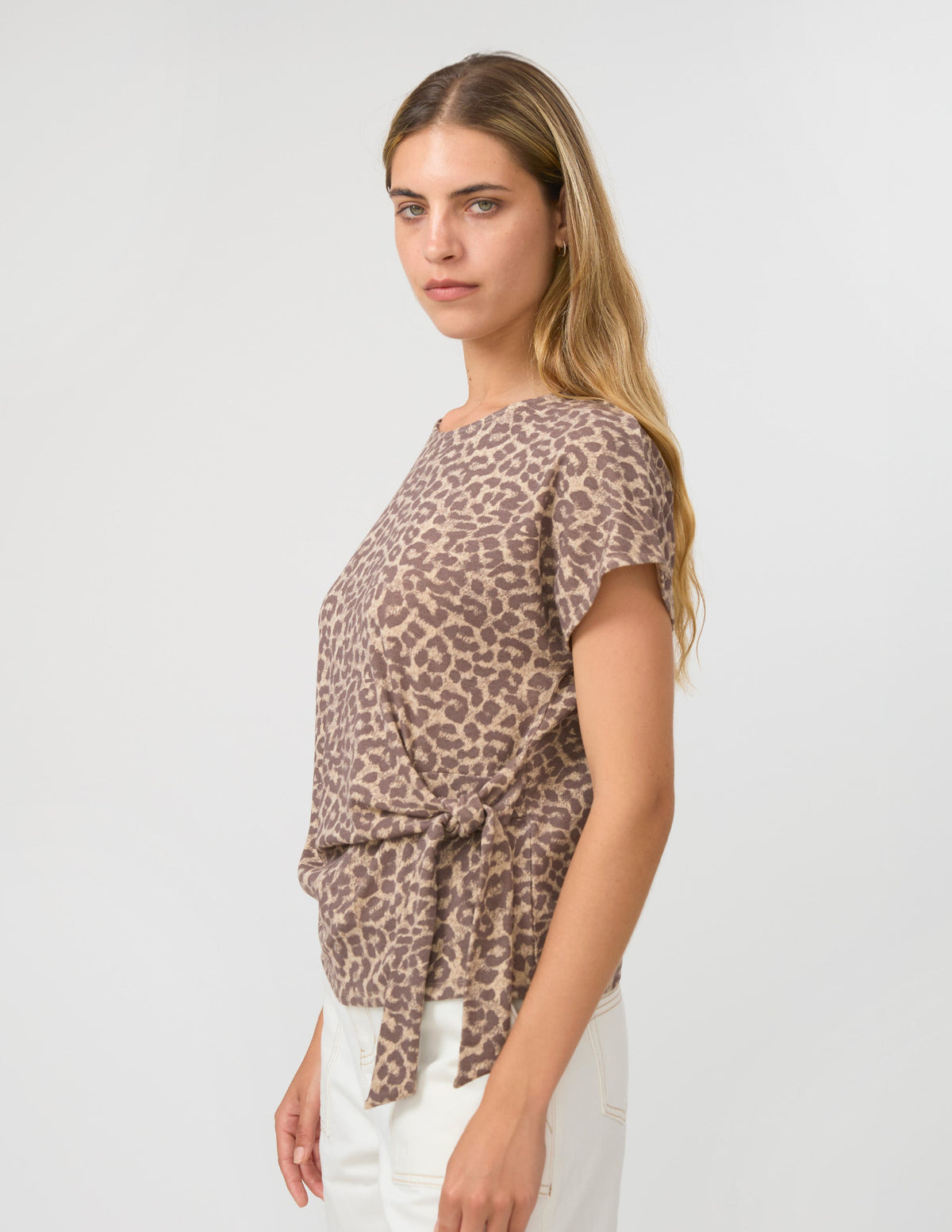 A woman modeling a short-sleeve leopard print top with a side-tie detail, paired with white pants, against a plain light gray studio background.