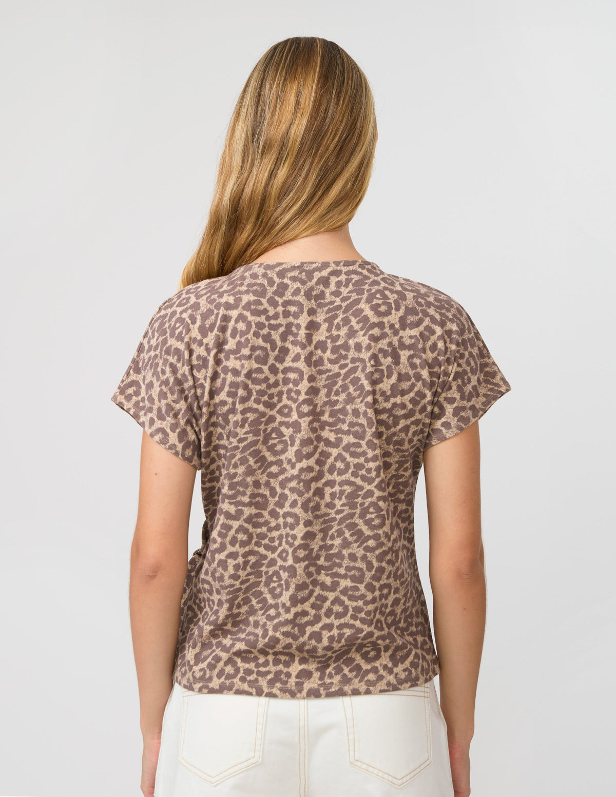 A woman viewed from behind wearing a short-sleeve crewneck t-shirt with a brown and beige leopard print, paired with white pants, against a light gray studio background.