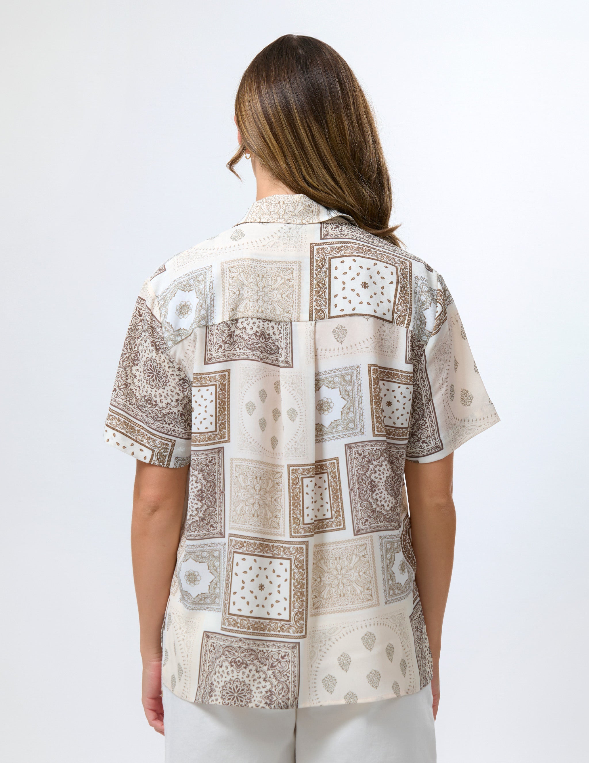 A woman viewed from the back wearing a short-sleeve button-up shirt with a beige, brown, and white patchwork print featuring geometric and floral motifs.