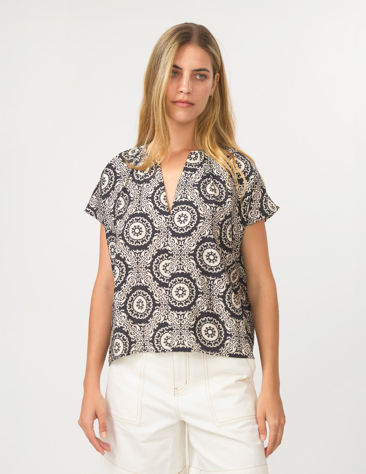 A woman modeling a short-sleeve button-up blouse with a black and off-white circular mandala pattern, paired with white shorts, against a plain light gray background.